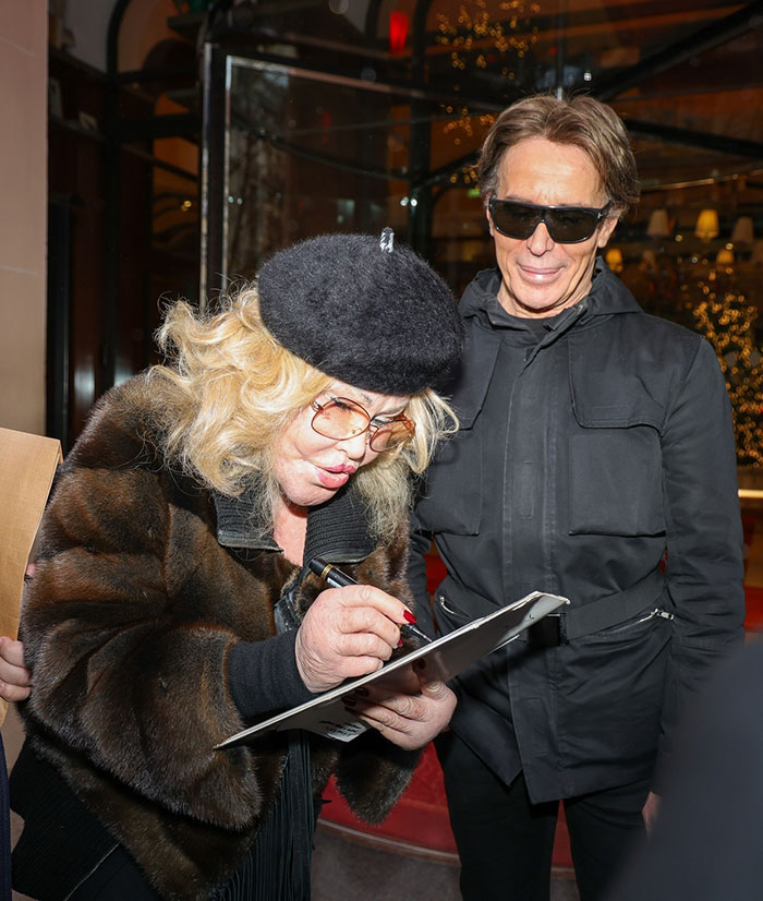 Jocelyn Wildenstein in Paris, wearing a fur coat and beret, with fianc&eacute; beside her, holding a clipboard.
