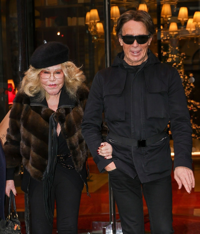 Jocelyn Wildenstein with smooth skin in Paris, wearing dark glasses and a fur coat, walking with a man in a black outfit.