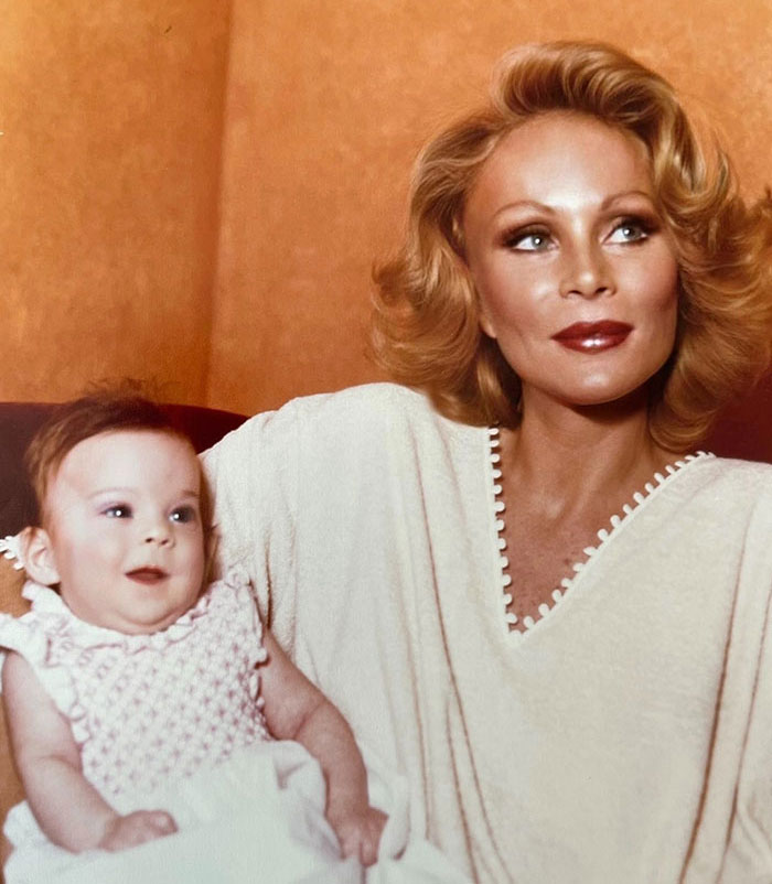 Jocelyn Wildenstein with smooth skin and styled hair holds a smiling baby, both in light clothing against a warm-toned background.