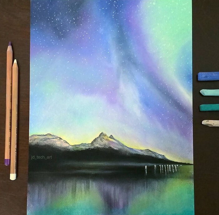 Hyper-realistic pencil drawing of a vibrant night sky over mountains and water, accompanied by colored pencils and pastels.