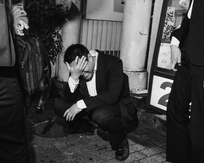 A man in a suit crouches in a Tokyo street, hand on head, illustrating the toll of fast-paced city life.
