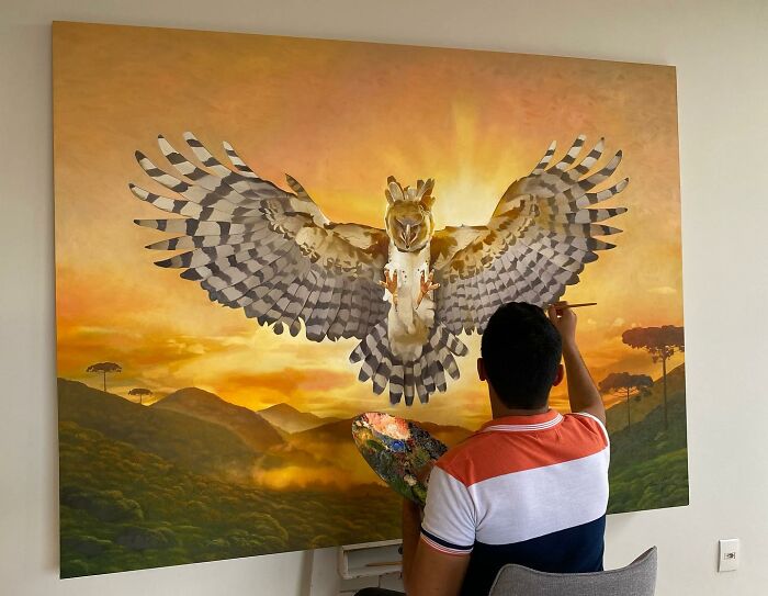 Brazilian artist painting a large feathered mural with vibrant sunset background.