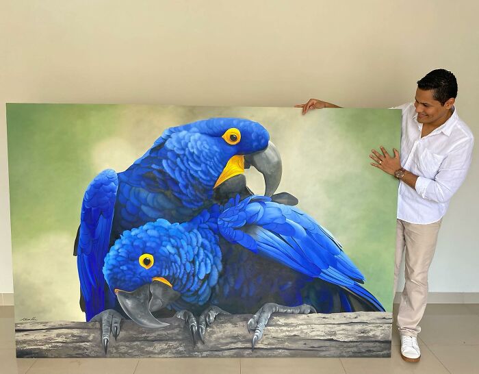 Petterson Silva showcasing vibrant feather art, featuring two blue parrots on a painted canvas.