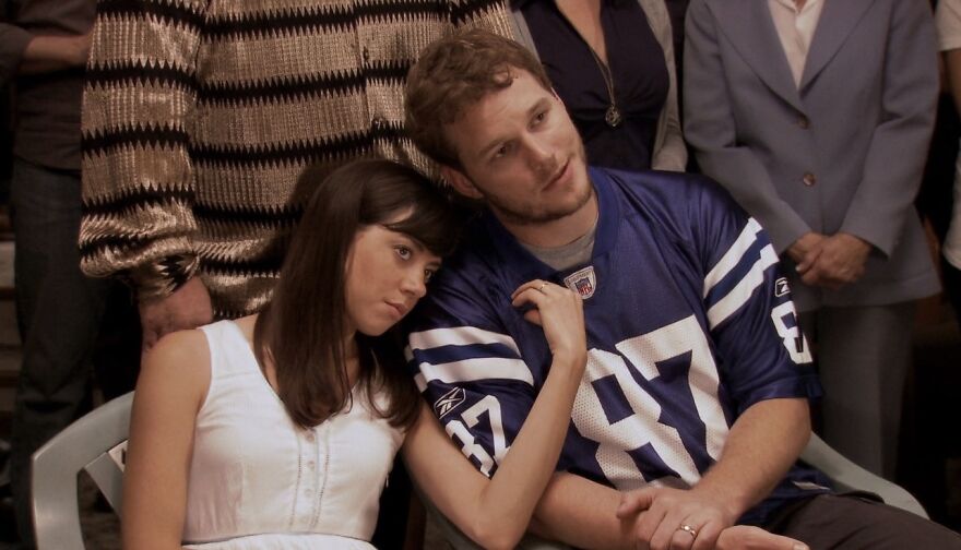 Couple sitting closely, woman in white and man in blue jersey, representing a movie relationship theme in Parks And Recreation.