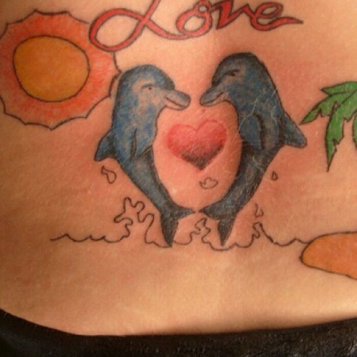 Two dolphins over a heart with a sun and palm tree in a tattoo, exemplifying a funny worst tattoo fail.