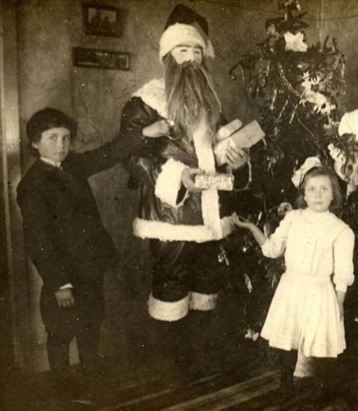 Children with a creepy Santa holding gifts beside a Christmas tree.