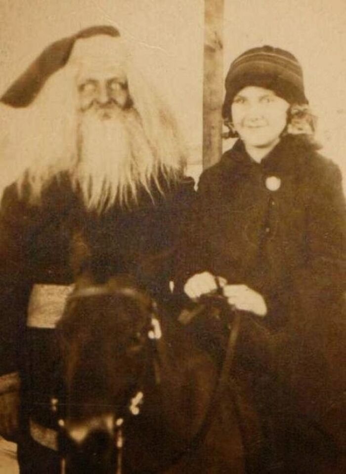Vintage photo of a creepy Santa with a long beard, standing beside a smiling child on a horse.