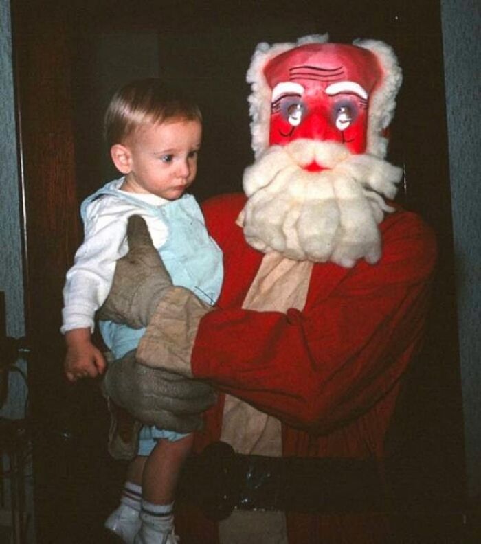 Child held by a creepy Santa with a red face and exaggerated features indoors.