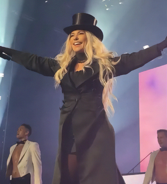 Shania Twain wearing a black top hat, performing energetically under bright lights.