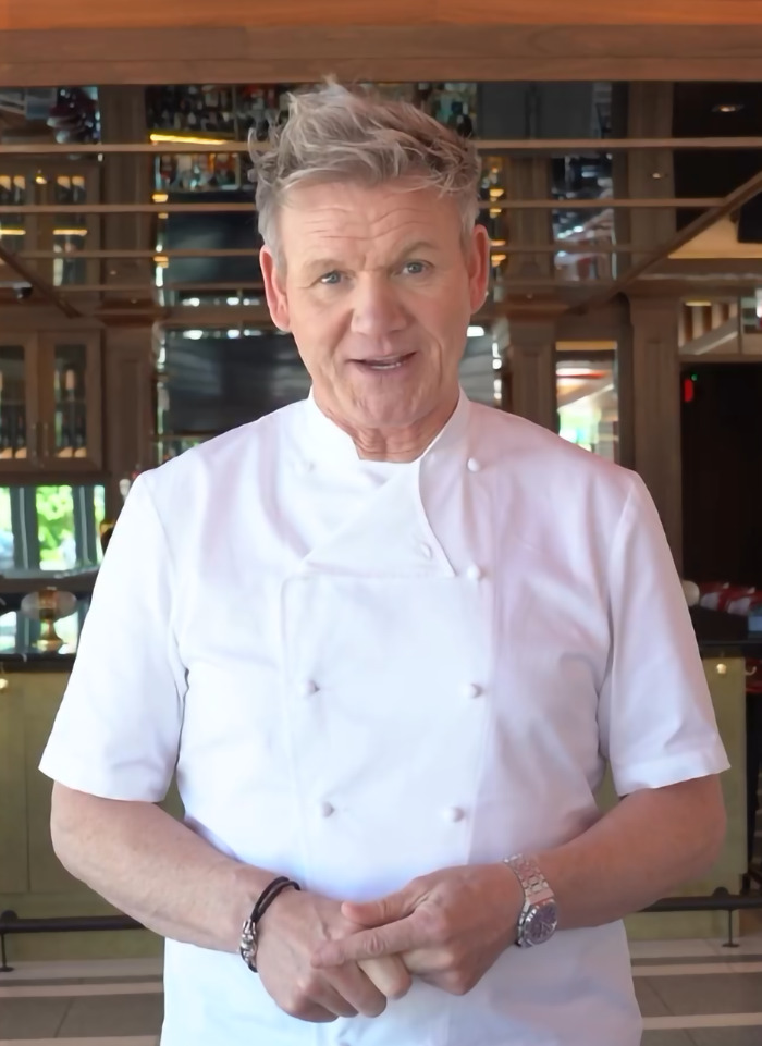 Chef Gordon Ramsay in a restaurant, wearing a white chef's coat, smiling.