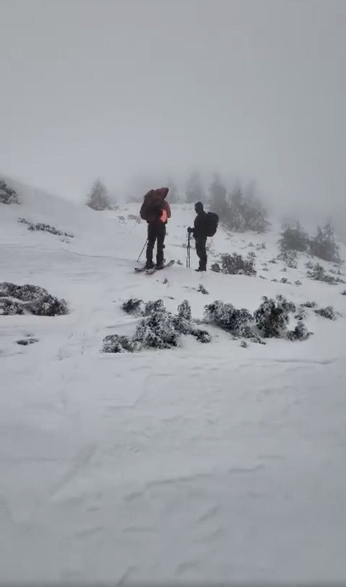 Two hikers on snowy mountain terrain, one with a backpack, in foggy conditions.