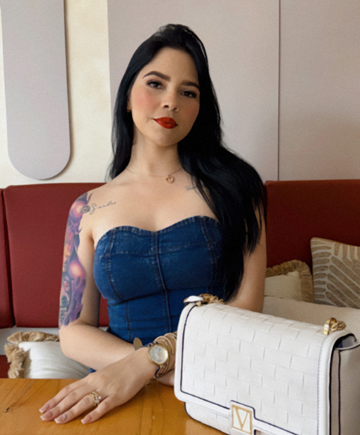 Nebrazkha in denim dress sitting at a table, with a white designer handbag.