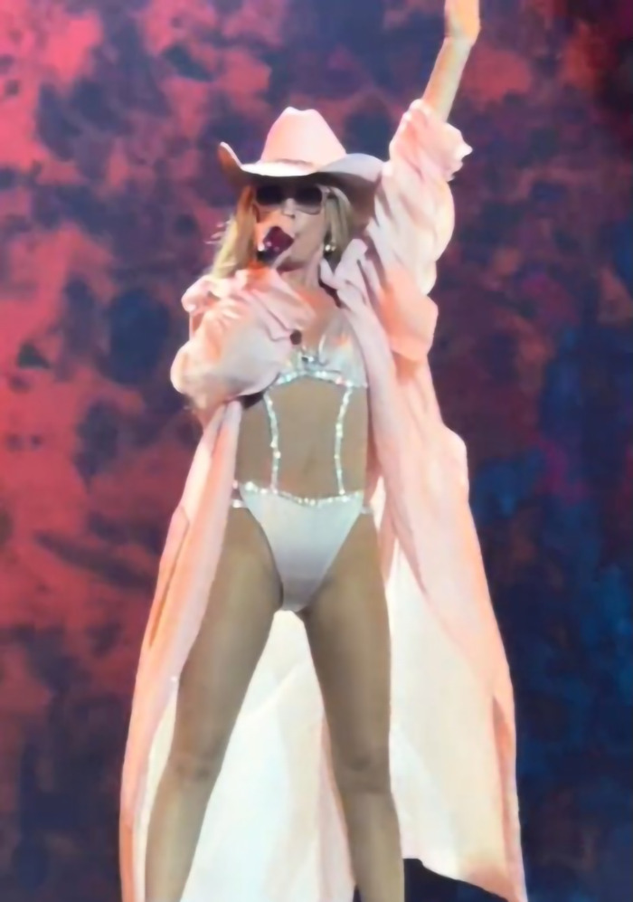 Shania Twain in rhinestone two-piece outfit on stage, raising one arm, wearing a hat and sunglasses.