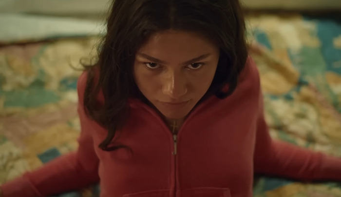 Zendaya in a scene, wearing a pink top, sitting on a patterned bedspread.