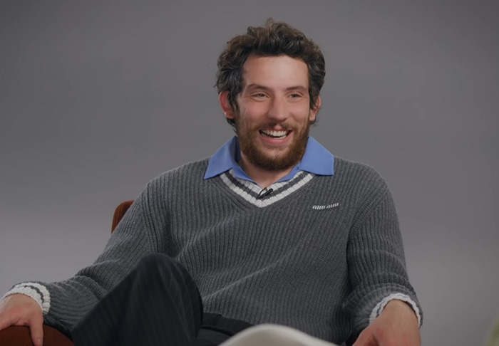 Josh O'Connor in a grey sweater and blue shirt smiles during an interview discussing Zendaya's X-rated scene.