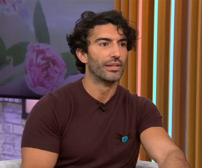 Justin Baldoni sitting on a talk show set, wearing a brown shirt, with floral backdrop and wooden paneling. Justin Baldoni sitting on a talk show set, wearing a brown shirt, with floral backdrop and wooden paneling.