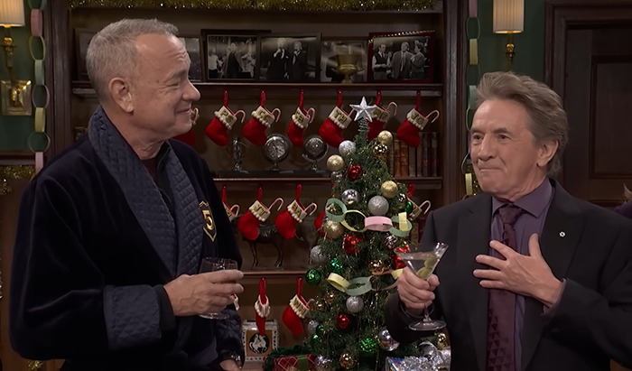 Tom Hanks on SNL, holding a drink, standing beside Martin Short near a Christmas tree.