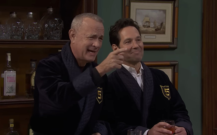Tom Hanks and Paul Rudd in dark robes on SNL set, one gesturing with hand, holding a drink.