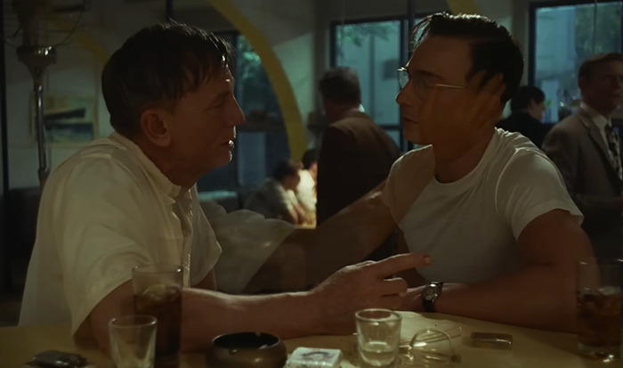 Daniel Craig sitting at a dimly lit bar engaged in an intense conversation, with drinks and a pack of cigarettes on the table on a scene from movie 'Queer'.