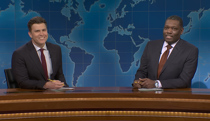 Colin Jost and co-host Michael Che at a news desk, smiling, with a world map backdrop.