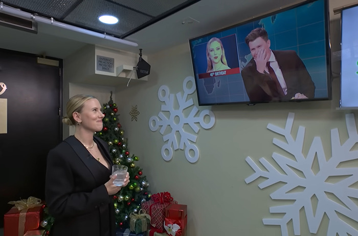 Scarlett Johansson smiles while watching Colin Jost on TV, with snowflake decorations and gifts in the background.