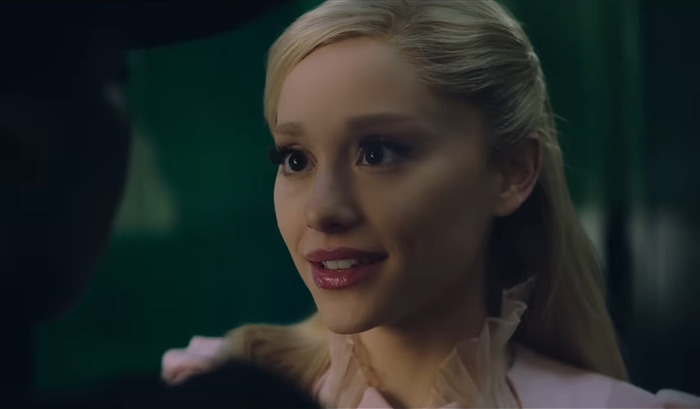 Ariana Grande as Glinda, facing a dark-clad person in a dimly lit setting from movie 'Wicked'. Ariana Grande as Glinda, facing a dark-clad person in a dimly lit setting from movie 'Wicked'.