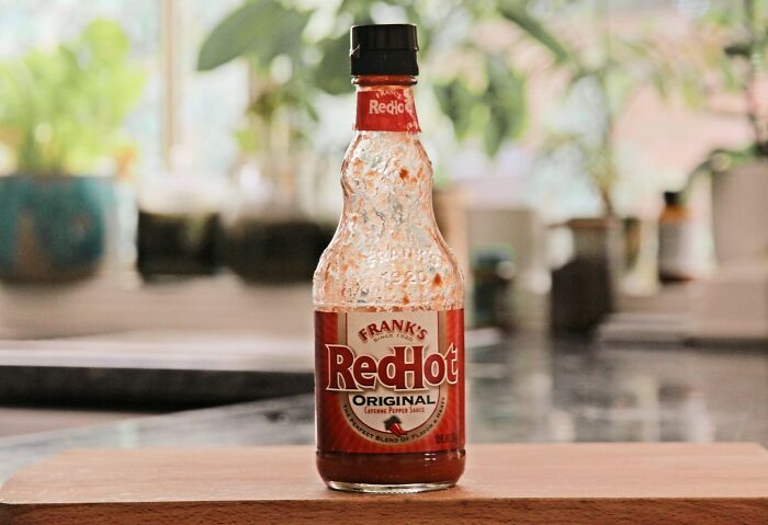 RedHot sauce bottle on a wooden table, surrounded by a blurred kitchen background.