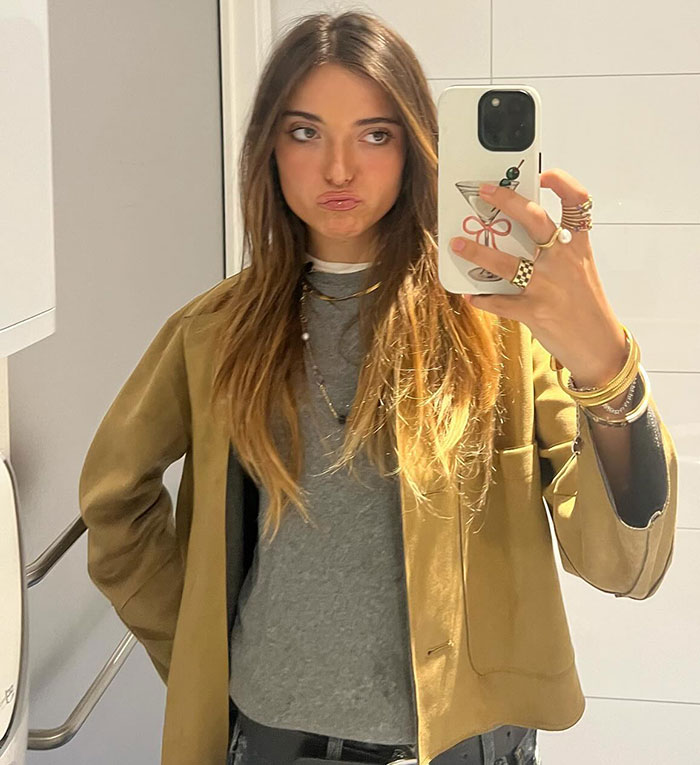 Woman taking a mirror selfie, wearing a brown jacket and gray shirt, with "Mean Girl" antics trending.