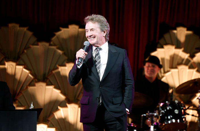 A smiling man in a suit holding a microphone on stage, related to Aquaman 2 cast event.