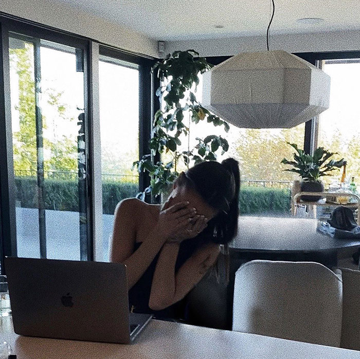 Woman at a table with hands over her face, sitting in front of a laptop in a bright room with plants.