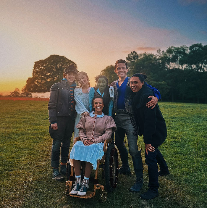 Cast members on set of "Wicked," standing together outdoors with a sunset in the background.