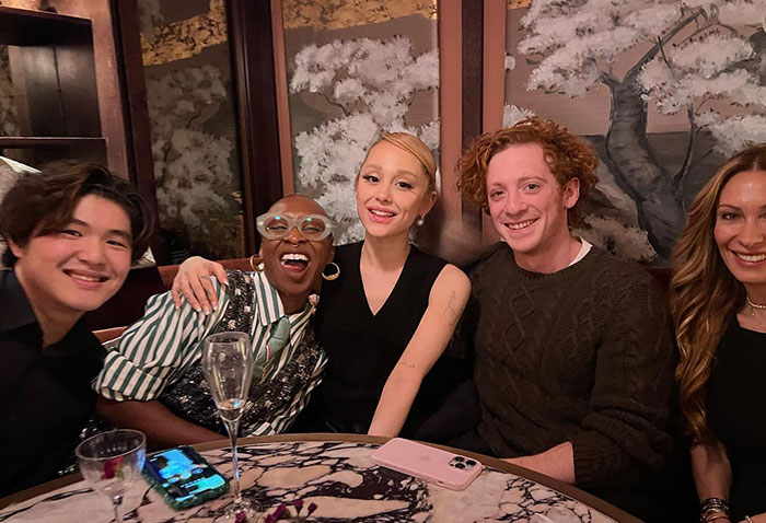 Group of friends smiling at a restaurant table, featuring Ariana Grande and Ethan Slater enjoying a night out.