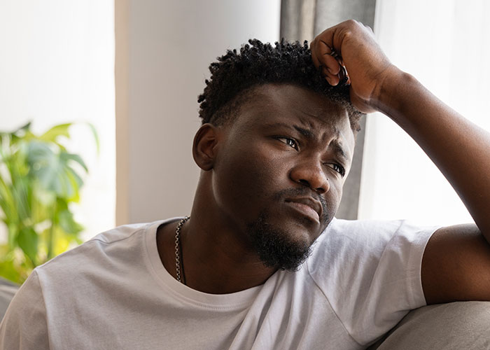 Man looking pensive, wearing a white shirt, sitting on a couch, light streaming in from a nearby window. Man looking pensive, wearing a white shirt, sitting on a couch, light streaming in from a nearby window.
