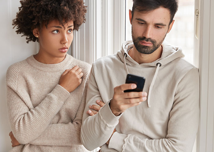 Couple standing by a window; the woman looks concerned as the man focused on his phone, highlighting relationship tensions. Couple standing by a window; the woman looks concerned as the man focused on his phone, highlighting relationship tensions.