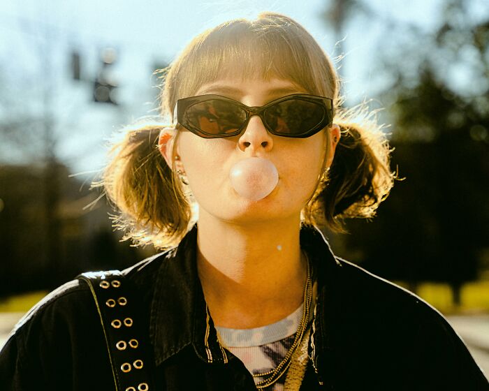 Person with sunglasses blowing bubble gum outdoors, demonstrating a bad-good-habit on a sunny day.