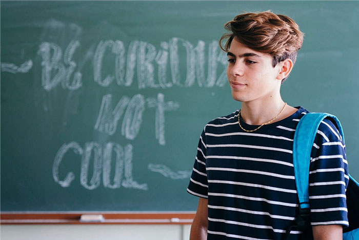 Student in striped shirt with backpack, standing in front of a chalkboard with unclear writing; terrible school presentation.