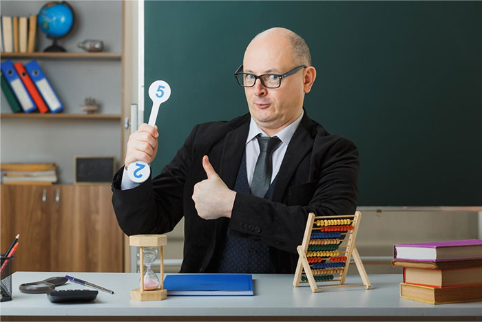 Teacher humorously holding number signs at desk with educational tools in front of blackboard.