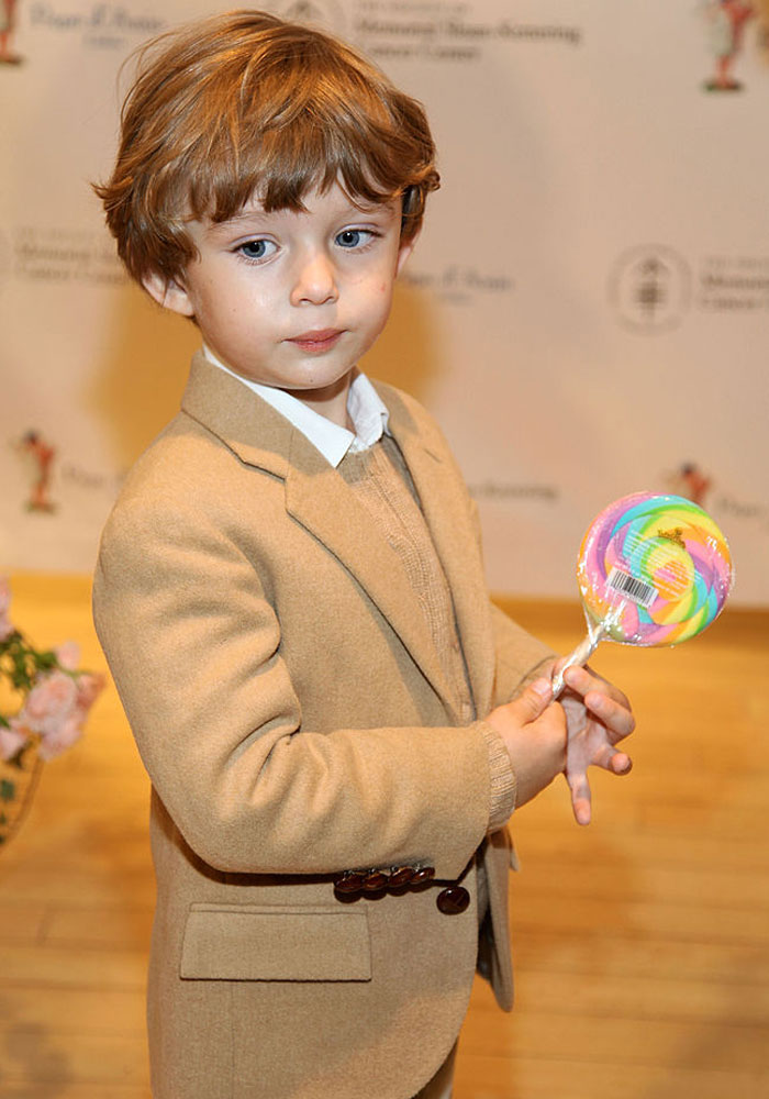 Young boy in a tan suit holding a colorful lollipop, with a thoughtful expression.