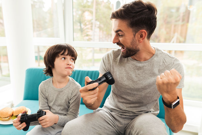 Man and boy playing video games, the man celebrating while the boy looks frustrated on a sofa, holding controllers. Man and boy playing video games, the man celebrating while the boy looks frustrated on a sofa, holding controllers.