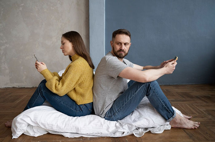 Young couple sitting back-to-back, looking at phones, symbolizing relationship regrets in their 20s.