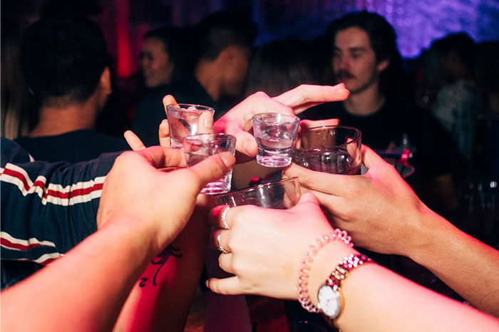 Group of friends clinking shot glasses at a lively party, representing a common regret from people's 20s.