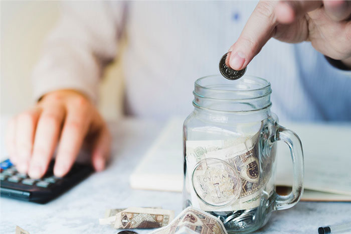 Person placing a coin in a jar, symbolizing financial regrets in people's 20s, with a calculator nearby.