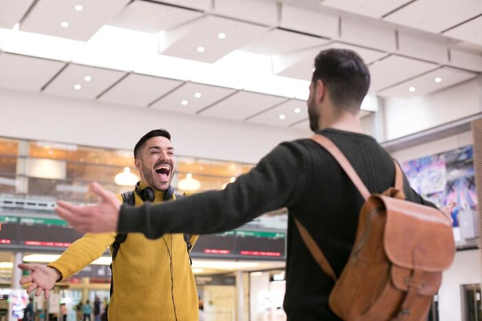 Two men joyfully greet each other at an indoor location, one wearing a brown backpack.