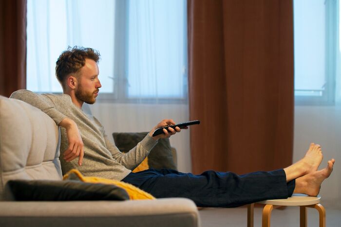 Man lounging on a couch, remote in hand, watching TV; represents a deadbeat freeloader visit.