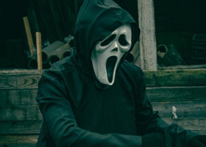 Person in a "Scream" mask and black cloak sitting outdoors, evoking a surprising home scene.
