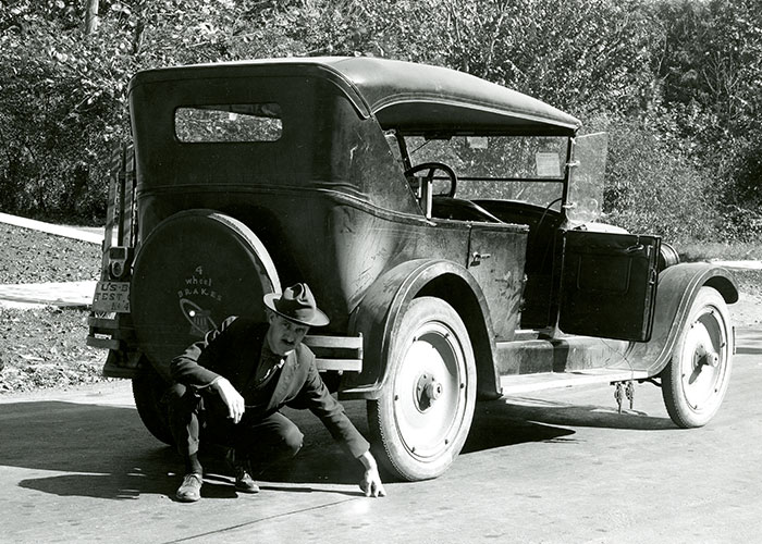 Vintage car with a person measuring something on the ground, evoking surprise in a home setting.
