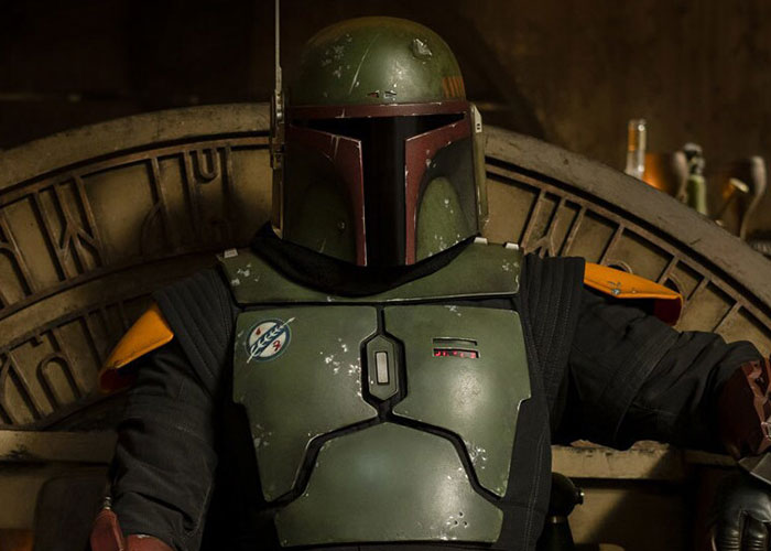 Person in green and brown armor sitting on a decorated throne, wearing a helmet.