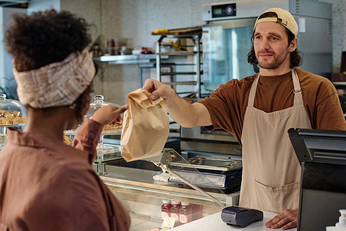 Employee in a cafe wearing an apron, handing a paper bag to a customer, illustrating reduced work hours.