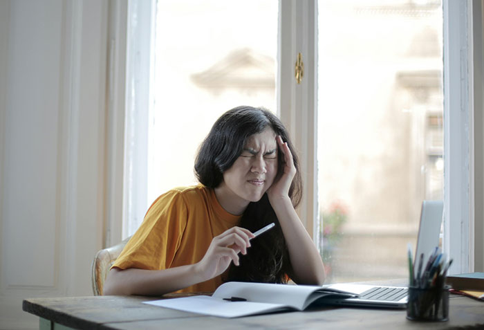 Woman in orange shirt stressed over wedding chores at desk with laptop and notebook.