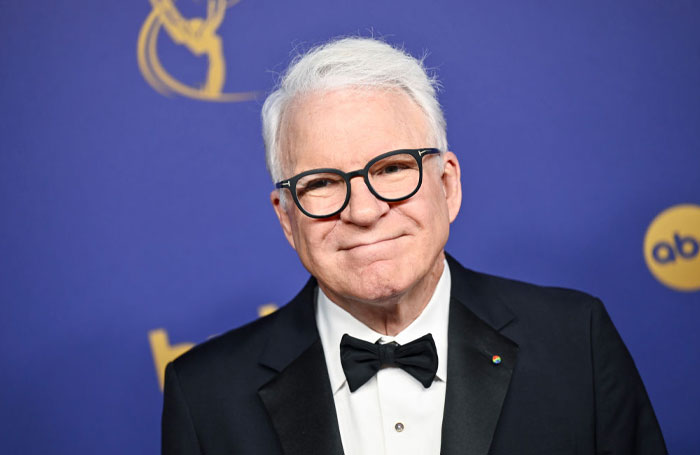 A cast member of Only Murders in the Building Season 4, in a tuxedo, smiling at an event with a purple backdrop.
